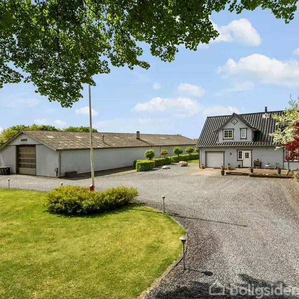 En grusvej fører til en hvid bygning ved siden af en grøn plæne og have. Der er også en stor garagebygning, omgivet af træer og buske under en blå himmel.