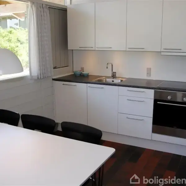 Hvidt køkken med integreret ovn og vask. Bord står ved vindue med udsigt til have. Lampe hænger over bordet, og der er sorte stole rundt om det.