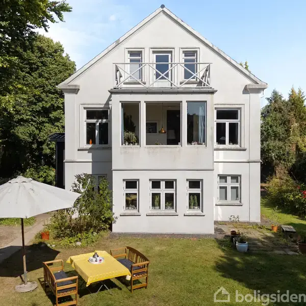 Et stort, hvidt hus med to etager står i en have. Foran huset er et havebord med gul dug, omgivet af fire stole, og en parasol.