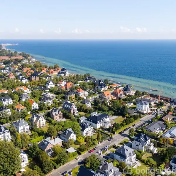 Kystnært boligområde med store villaer omgivet af grønne haver. Boligerne ligger tæt på hinanden langs en vej, med havudsigt og vand i baggrunden.