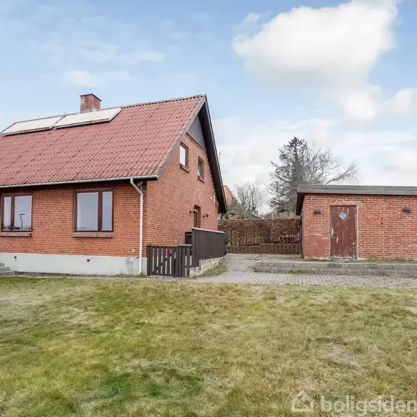 Rødt murstenshus med rødt tegltag står i en have med græsplæne. Til højre ses en separat murstensbygning med en trækontur. Himlen er klar med få skyer.