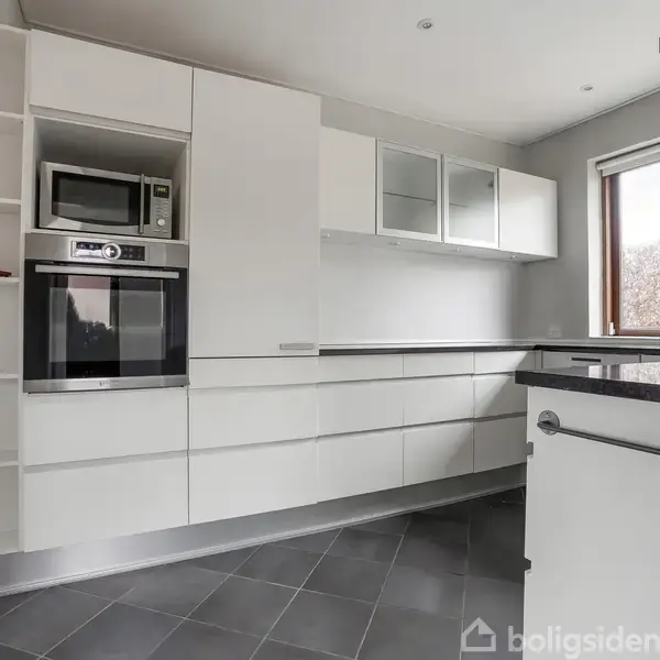 Et moderne køkken med hvide skabe og sort bordplade. Der er en indbygget ovn og mikrobølgeovn. Store vinduer giver udsigt til en hæk udenfor. Flisegulvet er gråt.