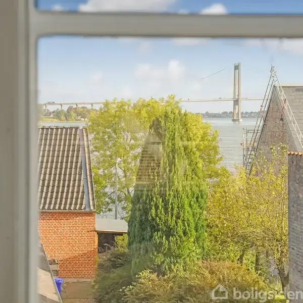 Et vindue viser en udsigt over hustage og træer mod en vandstrækning med en bro i baggrunden under en klar himmel.