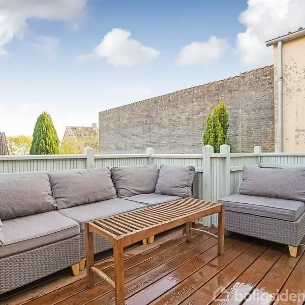 Grå sofa- og lænestolsæt står på en træterrasse med lavt hegn og planter, omgivet af nabobygninger. Himmel med skyer ses i baggrunden.