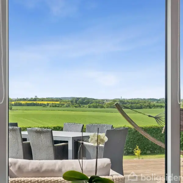 Glasdør med udsigt til terrasse med spisebord og stole, omgivet af grønne marker og blå himmel. En hængekøje fylder rummet ved siden af bordet, skaber afslappet stemning.