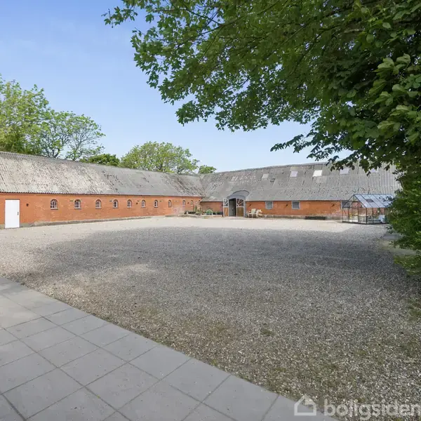 En rød bygning med en række små vinduer står stille omkranset af grønne træer og en stor grusbelagt gårdsplads. Der er en flisebelagt sti i forgrunden.