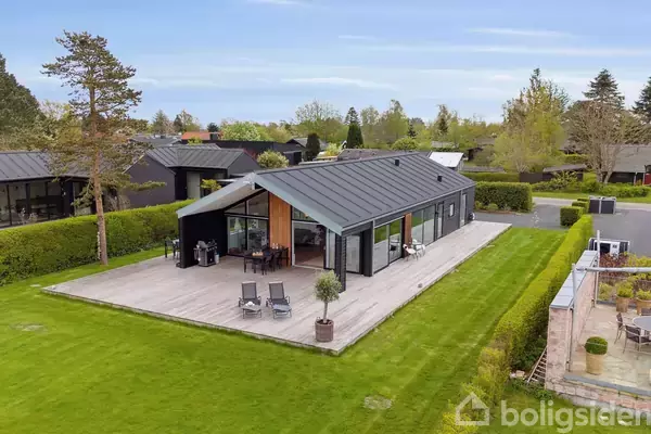 En moderne, sortmalet én-etagers villa står roligt med en rummelig træterrasse, udstyret med havemøbler. Omgivet af en velplejet have og placeret i et villakvarter med andre huse.