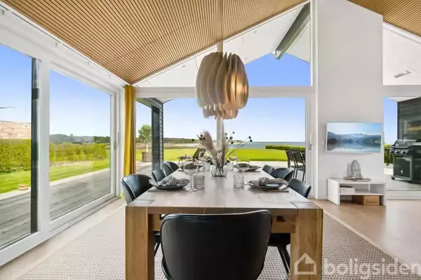 Spisebord med seks stole er dækket til et måltid, placeret i et moderne rum med store vinduer, der giver udsigt til en grøn have og blå himmel udenfor.