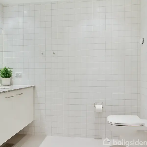 Toilet er monteret på en hvidkaklet væg, ved siden af en toiletpapirholder. Ved siden af står en vask med planter på en marmoroverflade. Rummet er lyst og minimalistisk.