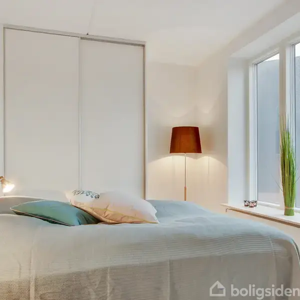 Soveværelse med en stor seng placeret mod et stort vindue, omgivet af flere puder. En lampe lyser fra hjørnet, og høje grønne planter står på vindueskarmen. Maleri hænger på væggen.