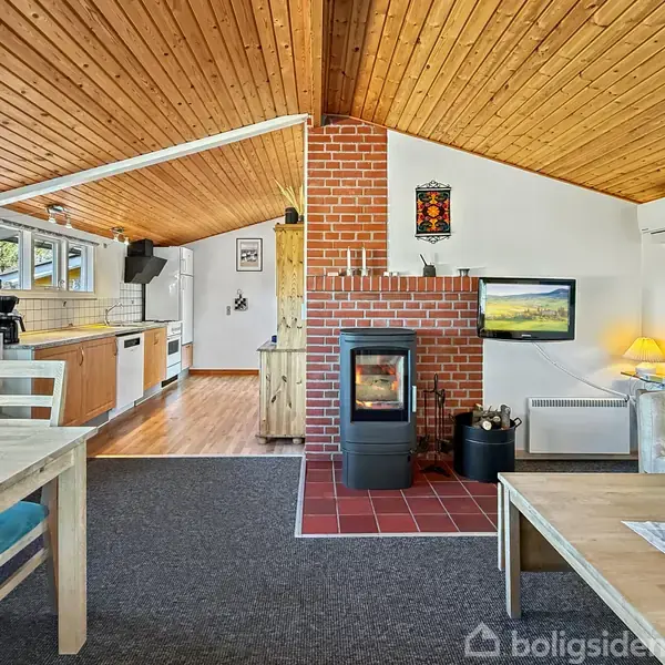 En brændeovn står ved en murstensvæg, opvarmende stue-spiseområde. Træloft og gulvlampe skaber en hyggelig atmosfære. Køkkenet ses i baggrunden til venstre med moderne apparater.