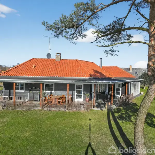 Et hus med rødt tag står på en grøn græsplæne. Huset har en stor veranda med træmøbler og er omgivet af træer og nabohuse i et villakvarter.