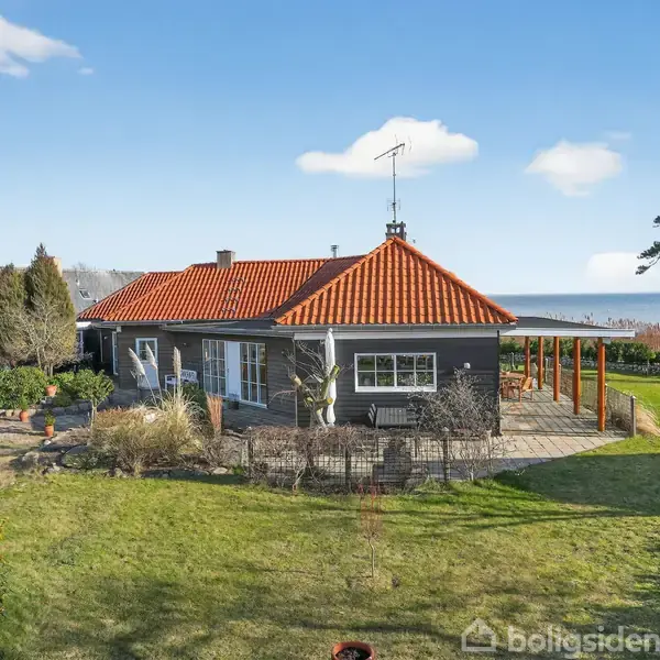 Hus med rødt tegltag står i en have. Huset har en stor terrasse og omgivende græsarealer. I baggrunden ses havet og nogle træer.