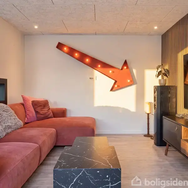 En rød sofa med puder står i en stue. En lysende pil peger nedad på væggen. Bord i sort marmorlook er foran sofaen. Reol med planter ses i højre side. Tegn med tekst "ANTIK" er synligt.