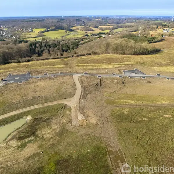 Anlægsplads med grusvej strækker sig gennem åbent landskab. Omgivet af marker og skove med en by og vindmøller i baggrunden under en blå himmel.