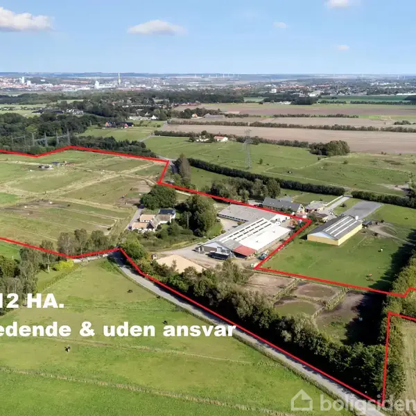 Indhegnet landområde med bygninger, marker og veje, set fra oven. Rød streg markerer området. Tekst: "Ca. 12 HA. Vejledende & uden ansvar." Landskabet omfatter marker og fjerntliggende by.