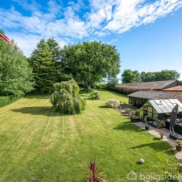 Flagstang med dansk flag står i en velplejet have med klippede plæner og træer. Ved siden af huset med terrasse og havemøbler. Skyfri himmel giver en solrig stemning.