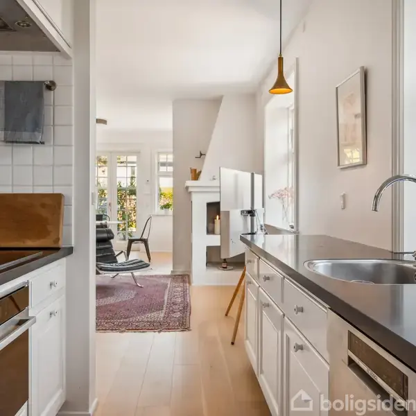 Et moderne køkken med hvidt interiør og sort bordplade er forbundet til en stue med pejs, lænestol og tæppe. Rummet er lyst med store vinduer.