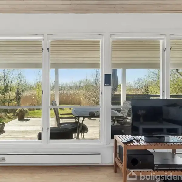 Et fjernsyn står på et bord, vendt mod en stor glasvæg. Udenfor ses en terrasse med borde og stole omgivet af grøn natur og træer i baggrunden.