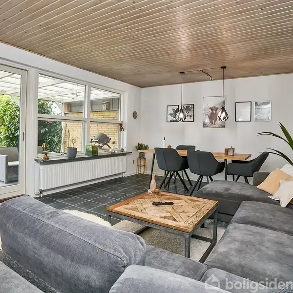 Stor grå sofa står i et moderne stueområde med træloft. Ved siden af sofabordet er der en spiseplads med sorte stole. Store vinduer giver udsigt til en udendørs terrasse.