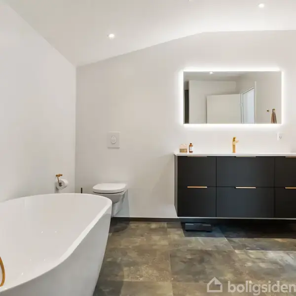 Et moderne badeværelse med fritstående hvidt badekar til venstre og en gylden bruser. En sort vaskeskab med spejl og belysning hænger på væggen, håndklæde til højre.