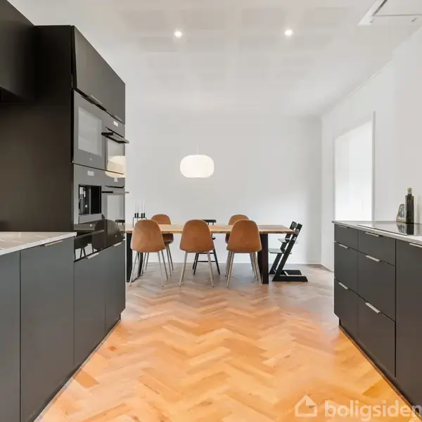 Sort køkken med moderne skabe på hver side, fører til spisebord med fem træstole under en minimalistisk lampe. Gulvet har sildebensparket. Rummet er lyst og rummeligt.