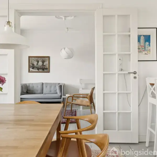 Blomster i en vase står på et træbord i et lyst rum. I baggrunden er der en grå sofa, hvide døre, vægkunst og træstole. Hjemlig atmosfære.