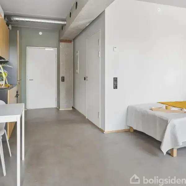 En minimalistisk lejlighed med en enkeltseng dækket af hvidt sengetøj og et gult tæppe. Til venstre står et lille spisebord med vaser og planter, der skaber en lys og indbydende atmosfære.