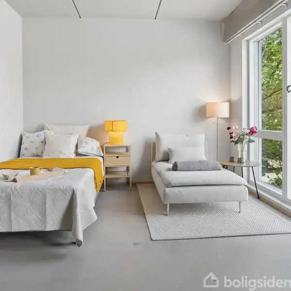 Dobbeltseng og chaiselong står ved siden af hinanden i et lyst værelse. Ved siden af senge er et natbord med lampe og en stor vindue bemærkes i baggrunden med udsigt til træer.