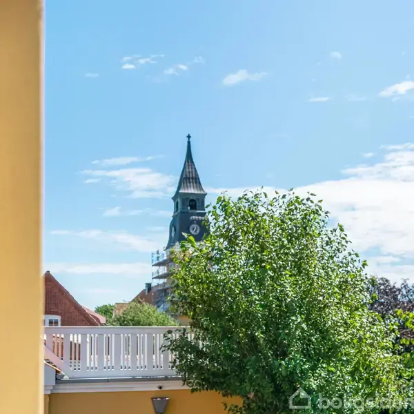 Et kirketårn med ur ses i baggrunden over en mur og grønne træer under en blå himmel med lette skyer.