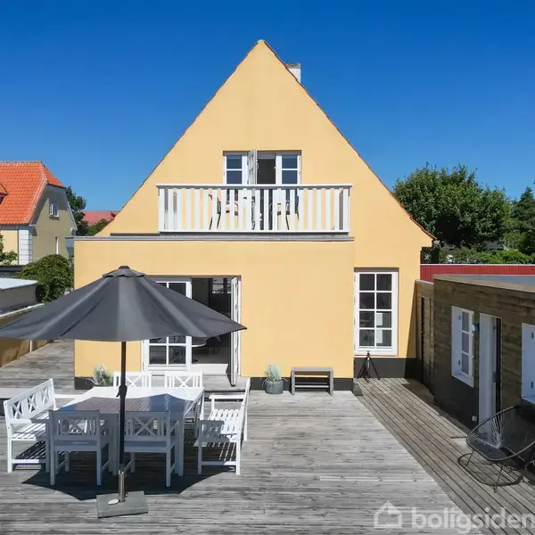 Gulmalet hus med trekantet gavl og balkon foran en stor træterrasse. Terrassebord og stole står under en parasol. Omkring ses andre hustage og grønne træer under en klar blå himmel.