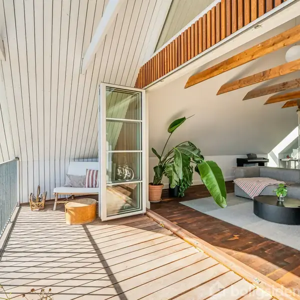En rummelig, lys stue med træbjælker i loftet åbner op til en altan med glasdør. En stor plante står ved siden af en moderne sofa, hvor et tæppe ligger. På altanen er der en lille sofa og bord med udsigt.