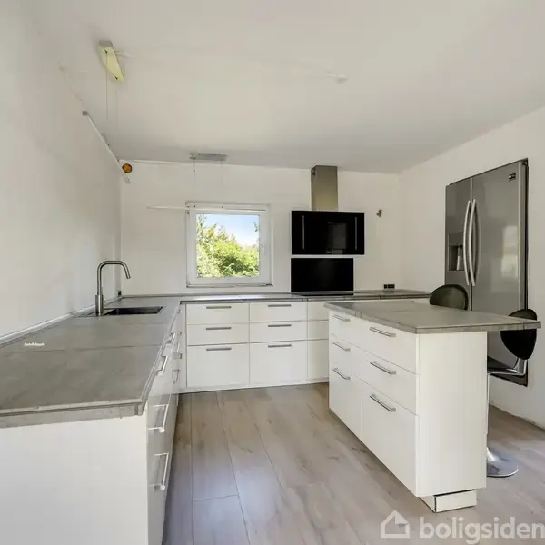Et moderne køkken med hvide skabe, en grå bordplade og en central ø. En stor køleskab står til højre. Gennem vinduet ses grønne træer udenfor.