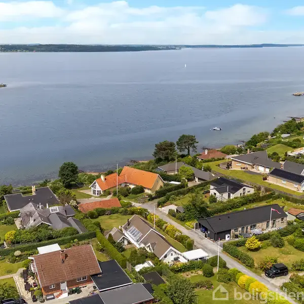 Adskillige huse med haver ligger langs en kystlinje. Handlinger er fraværende, mens konteksten viser en rolig vandudsigt med en klar himmel.