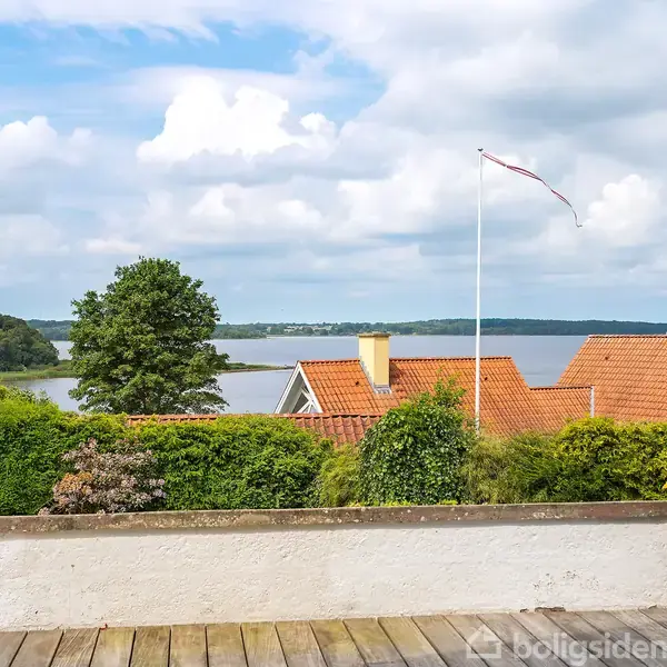 Terrasse med trægulv, omkranset af grønne buske, udsigt til røde tegltage og havudsigt i baggrunden under en overskyet himmel. En flagstang med hvidt flag står synligt.
