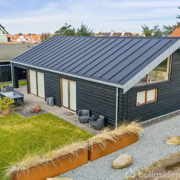 Hus med sort træbeklædning og mørkt tag står i en velplejet have med græsplæne og terrasse. Grusstier og dekorative sten omgiver huset i et beboelseskvarter med lav bebyggelse.