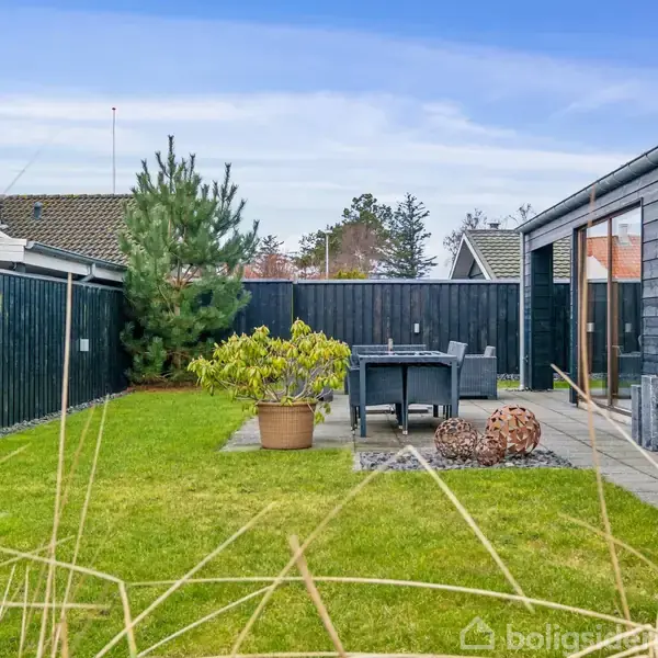 Have og terrasse møbleret med bord og stole. Omkranset af sortmalet hegn og græsplæne. Træbeklædt hus i baggrunden. Terrasse dekoreret med planter og kugler.