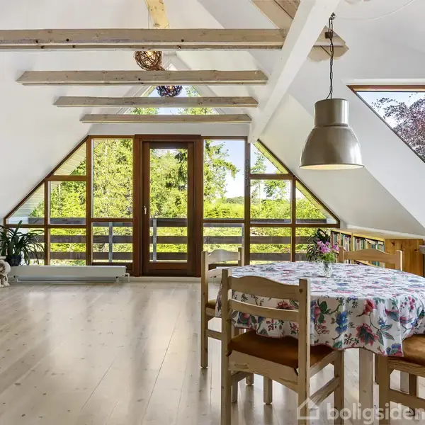 Et spisebord med en blomstret dug står ved vinduet. Rummet har synlige loftsbjælker, store vinduer med udsigt til grønne træer og en bogreol langs væggen.