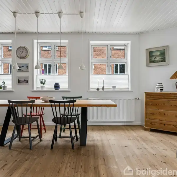 Spisebord med fire stole står i et lyst rum. Over bordet hænger lamper, mens væggen har kunst og ure. Skænk med lampe står ved siden af vinduer.