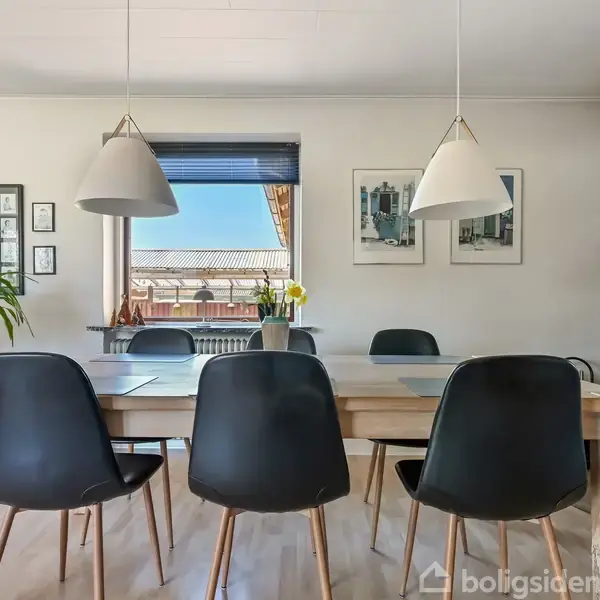 Et spisebord omgivet af sorte stole står i rummet. To lamper hænger over bordet med en brændeovn i hjørnet. Et vindue med blå persienner og billeder pryder væggen.