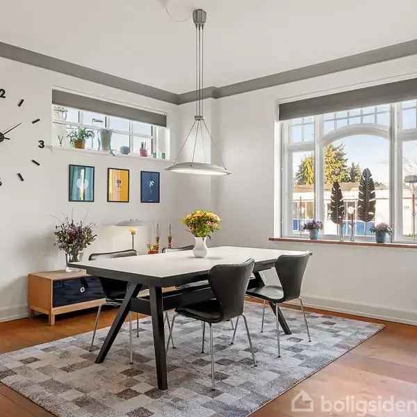 Et spisebord med fire stole står midt i et lyst rum. En stor vægur hænger på væggen. Væggene er dekoreret med billeder, og store vinduer slipper lys ind.