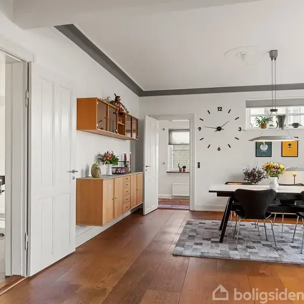 Spisestue med sort bord og stole, omkranset af en stor væg-ur, planter og kunst på væggen. Trægulv og vinduer bringer lys ind. Dør til tilstødende rum ses.