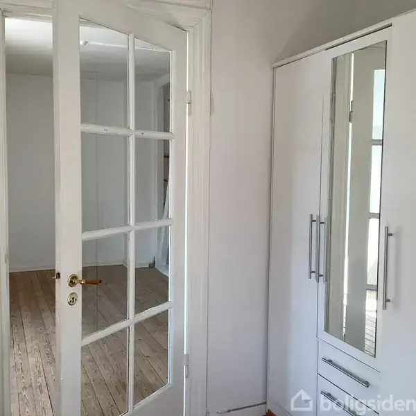 En hvid glasdør står åben og leder ind til et rum med træparketgulv. Til højre er der en garderobe med spejldøre. Rummet er lyst og enkelt.