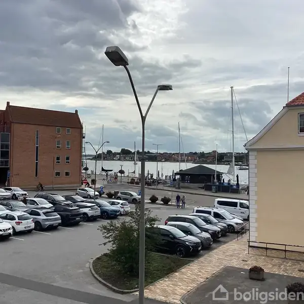En parkeringsplads med mange biler holdende stille foran murstens- og gule bygninger. I baggrunden ses en havnefront med både og en grå, skyet himmel.