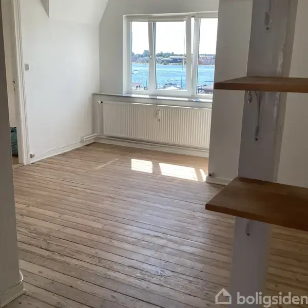 Et lyst, tomt rum med trægulv. Et stort vindue lukker naturligt lys ind, og der er udsigt over vandet. En radiator er placeret under vinduet.
