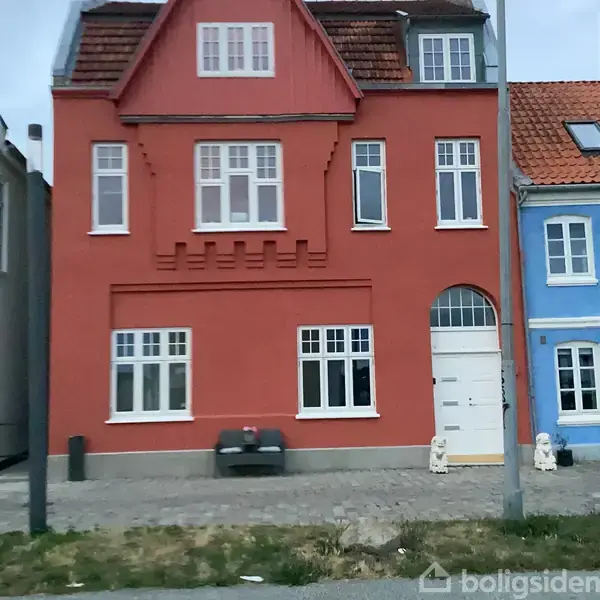 Et rødt to-etagers hus med hvide vinduer står ved siden af et blåt hus. Foran huset er en bænk og to stenskulpturer.