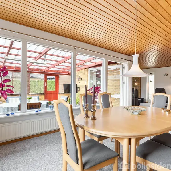 Rundt spisebord med fire stole står i en lys stue. Til venstre ses store vinduer med udsigt til en overdækket terrasse. Lyse trævægge og loft skaber en hyggelig atmosfære.