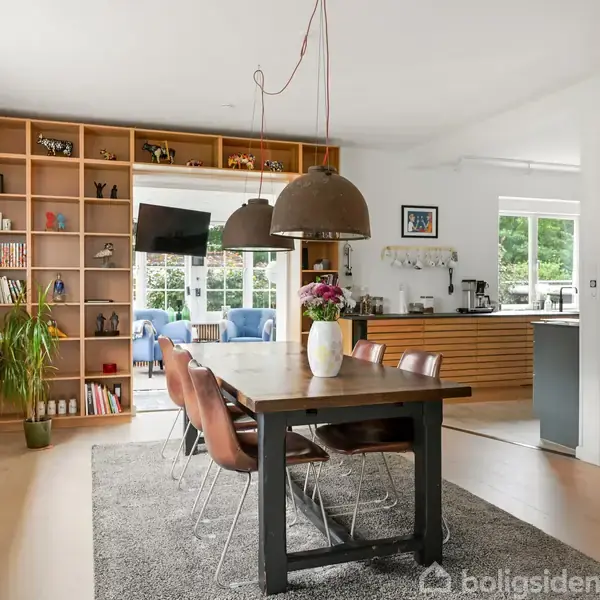 Et spisebord står med seks stole i et lyst rum. I baggrunden er en træbogreol fyldt med bøger og dekorationer. Et åbent køkken ses til højre.