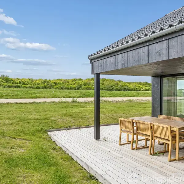 Et træterrassebord og stole står på en delvist overdækket terrasse ved et moderne træhus. Baggrunden er en grøn have med en åben himmel.