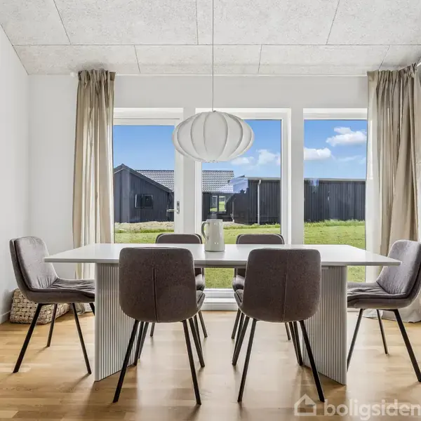 Et moderne spisebord står med seks stole rundt om det i et lyst og minimalistisk rum med store vinduer, der giver udsigt til en grøn græsplæne og et sort hus.